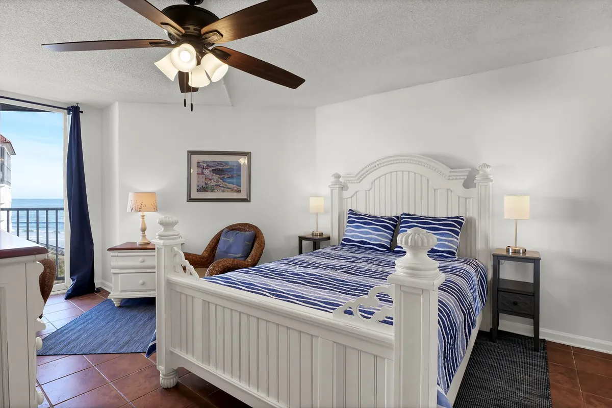 A tranquil master bedroom featuring a plush queen bed with blue striped bedding, a window offering a view of the ocean, a decorative lamp on the bedside table, and a woven chair positioned in a cozy corner. Natural light filters through the window, creating a soothing atmosphere ideal for relaxation.