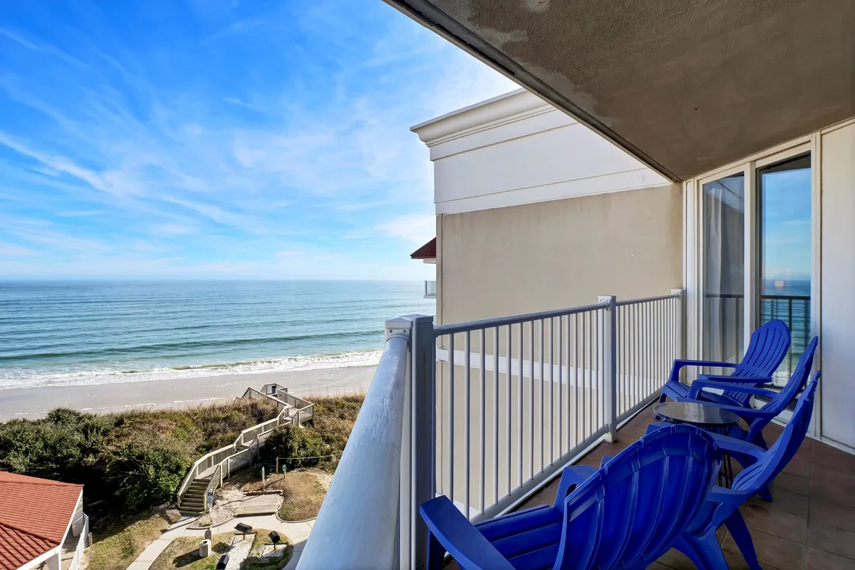 A serene ocean-view balcony featuring two comfortable chairs overlooking a sweeping seascape. The balcony is bordered by glass panels, offering unobstructed views of the sunrise, the gentle waves, and a pathway leading directly to the sandy beach below.