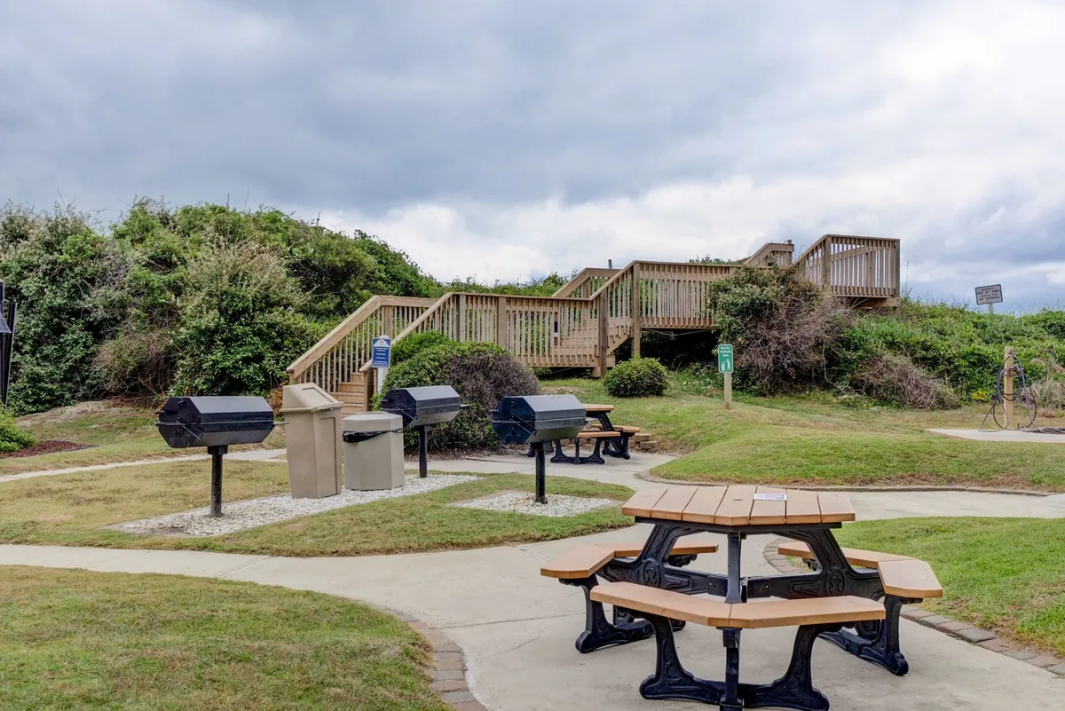 An outdoor picnic and barbecue area with sturdy tables and built-in barbecue grills, under the open sky. The setup suggests space for socializing, sunset meals, and family gatherings just steps from the resort's main buildings.