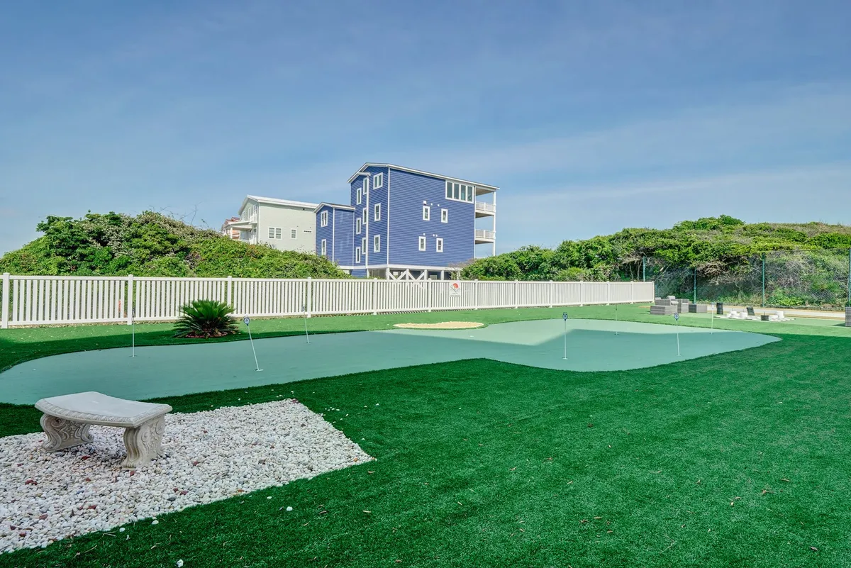This exterior image features an artificial putting green, a stone bench for relaxing, and a fenced yard next to a blue house. The scene is sun-drenched and inviting, suggesting both individual and group activities under open skies.