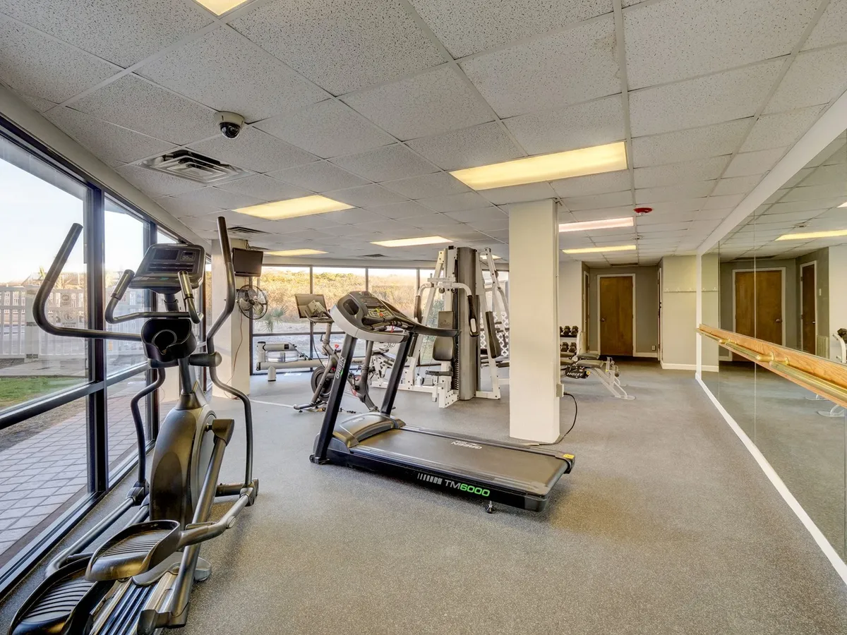 A bright, modern fitness center showcasing a treadmill, elliptical machine, weightlifting equipment, and large windows that let in natural light. The view outside hints at lush surroundings, and the space feels inviting and active.