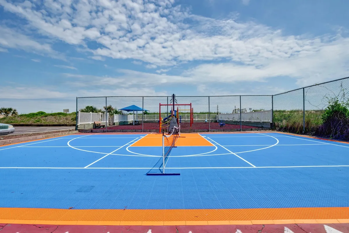 A colorful, multi-use sports court with a tennis net and playground equipment framed by lush greenery. The image hints at tennis, basketball, and family play, capturing the playful, active side of the property.