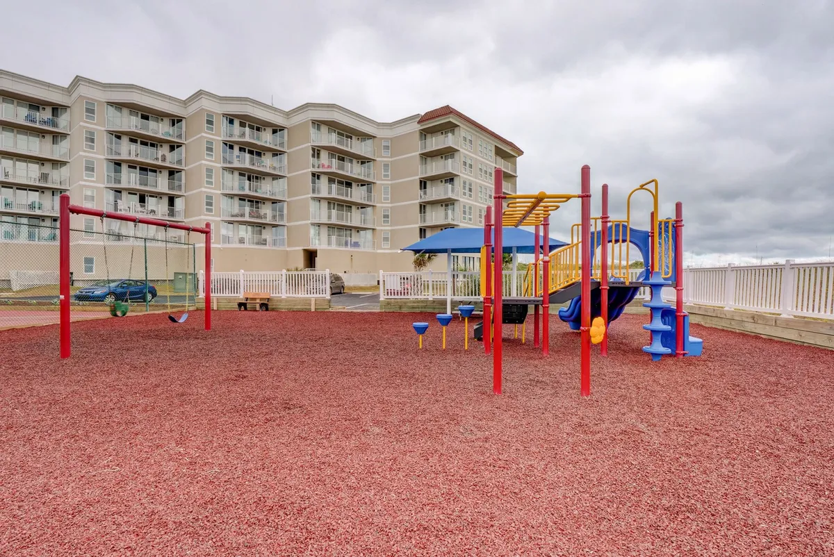 A vibrant playground featuring modern play equipment and a swing set, set atop safe red rubber flooring with a residential building in the backdrop. The cheerful environment is designed for children and evokes the sounds of laughter and play.