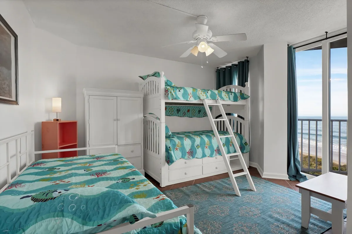 A cheerful second bedroom with twin bunk beds and a trundle, adorned in colorful bedding. Large windows overlook the ocean, and playful decor elements create an inviting, family-friendly space tailored for children or extra guests.