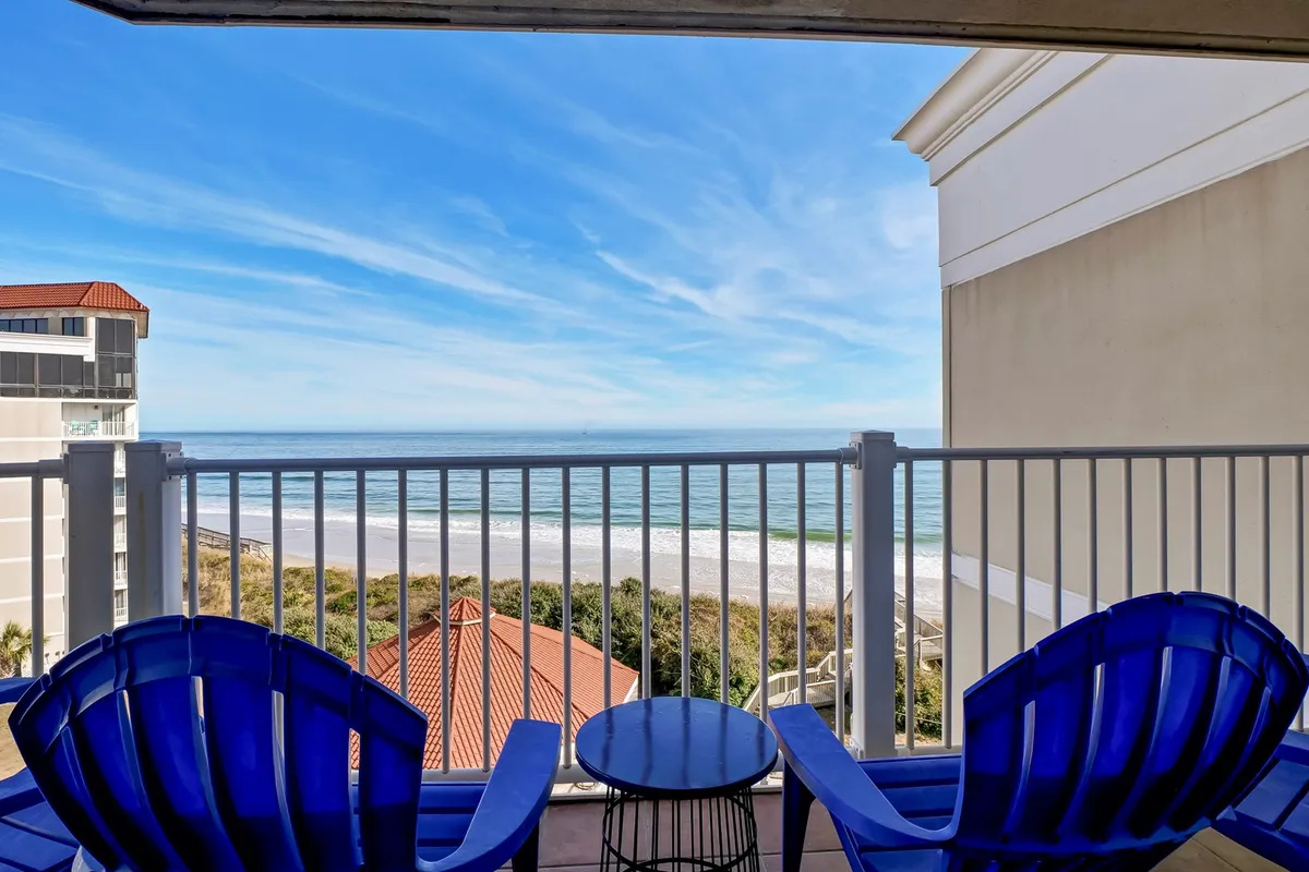 Two blue Adirondack chairs and a small round table sit on a balcony overlooking the beach and Atlantic Ocean, with the distant horizon visible. The scene suggests morning coffee or sunset relaxation amidst the fresh sea breeze.