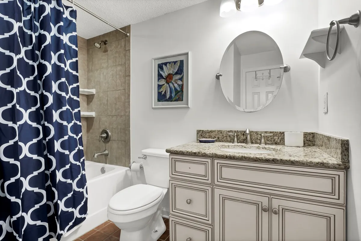 An inviting bathroom featuring a granite countertop, a blue patterned shower curtain, framed floral art, and a large mirror. The space feels bright and fresh, ideal for relaxing baths or preparing for the day.