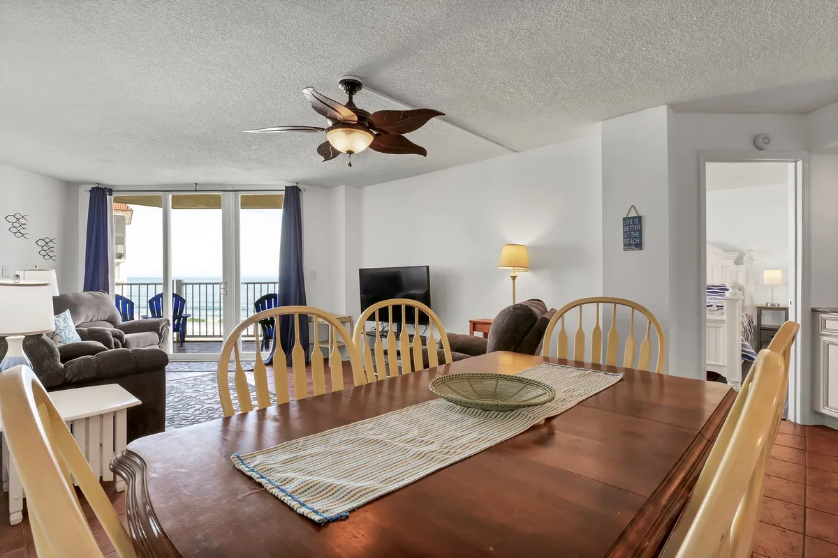 This image presents an inviting interior space featuring a dining table, cozy sofa, and ceiling fan, all oriented toward large windows framing an expansive view of the ocean. The tranquil ambiance is enhanced by soft natural light and the seamless flow between the seating and dining areas, with the balcony visible just beyond.